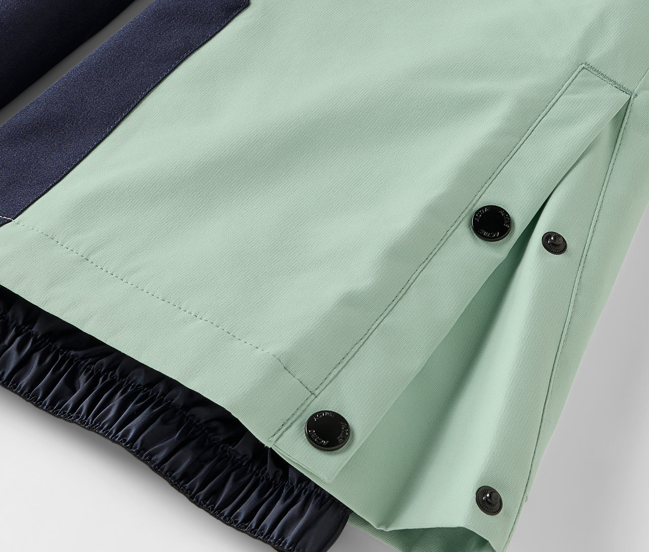 Detal jasnych, zielonych spodni z zapięciem na guziki i elastycznym obszyciem.