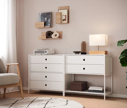 Dwie białe komody stoją obok siebie, na nich lampa, wazon i radio. Na ścianie wiszą półki.