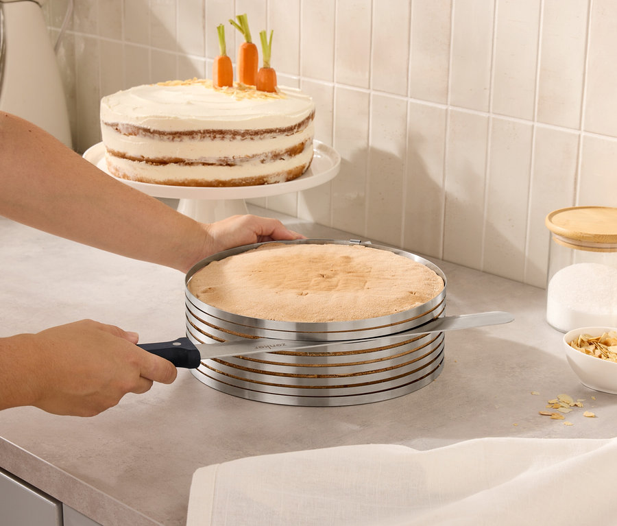 Osoba kroi ciasto za pomocą regulowanego krajalnicy do ciast. W tle znajduje się ciasto z marchewką i słoiki z pokrywkami.