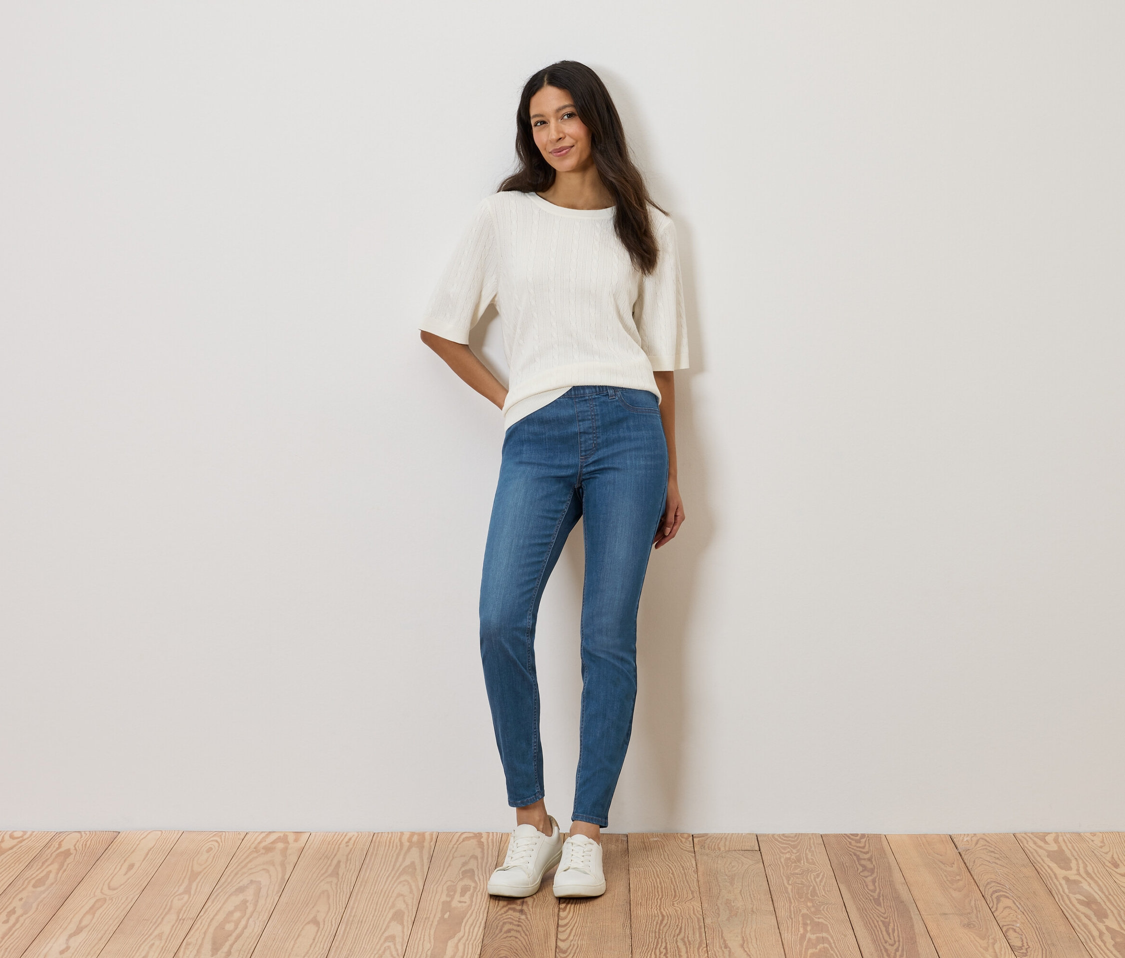 Kobieta stoi na tle ściany, ubrana w białą bluzkę, niebieskie dżinsy i białe buty.