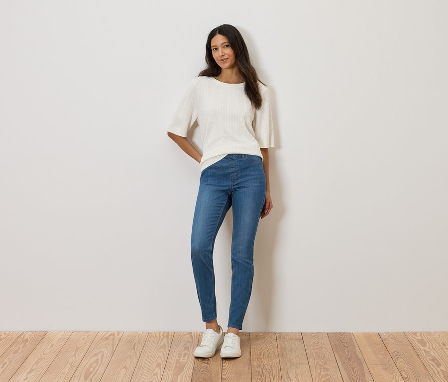 Kobieta stoi na tle ściany, ubrana w białą bluzkę, niebieskie dżinsy i białe buty.