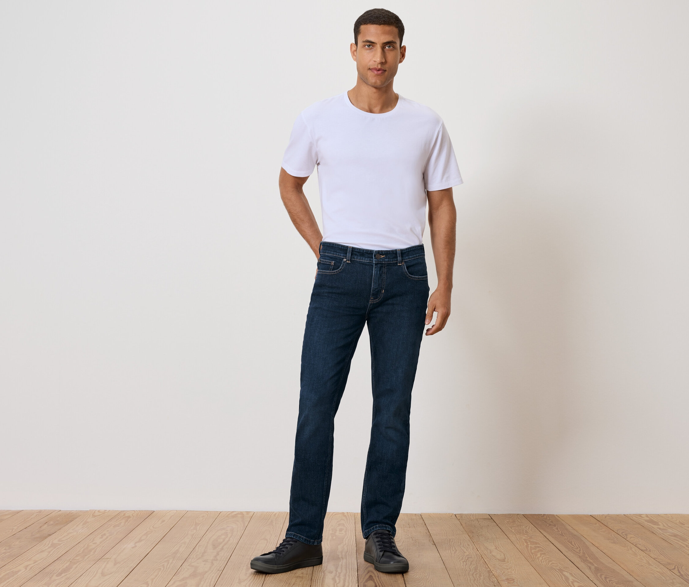 Mężczyzna stoi z ręką w kieszeni, ubrany w białą koszulkę, dżinsy i czarne buty.