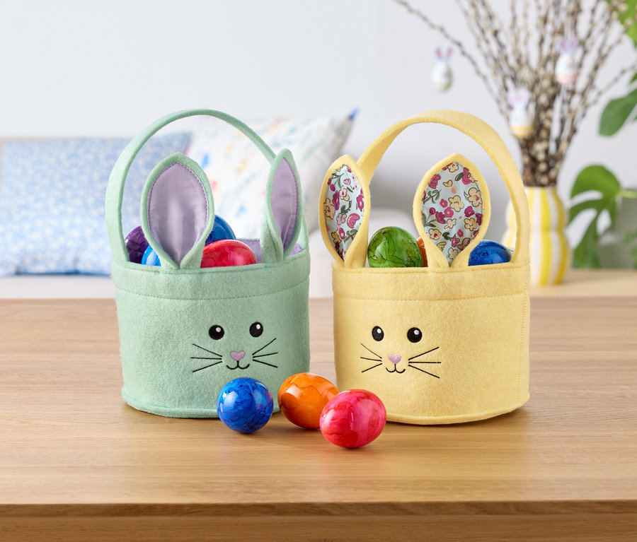 Dwa koszyczki wielkanocne w kształcie króliczka, jeden miętowo-zielony i jeden żółty, wypełnione kolorowymi jajkami, stoją na stole.