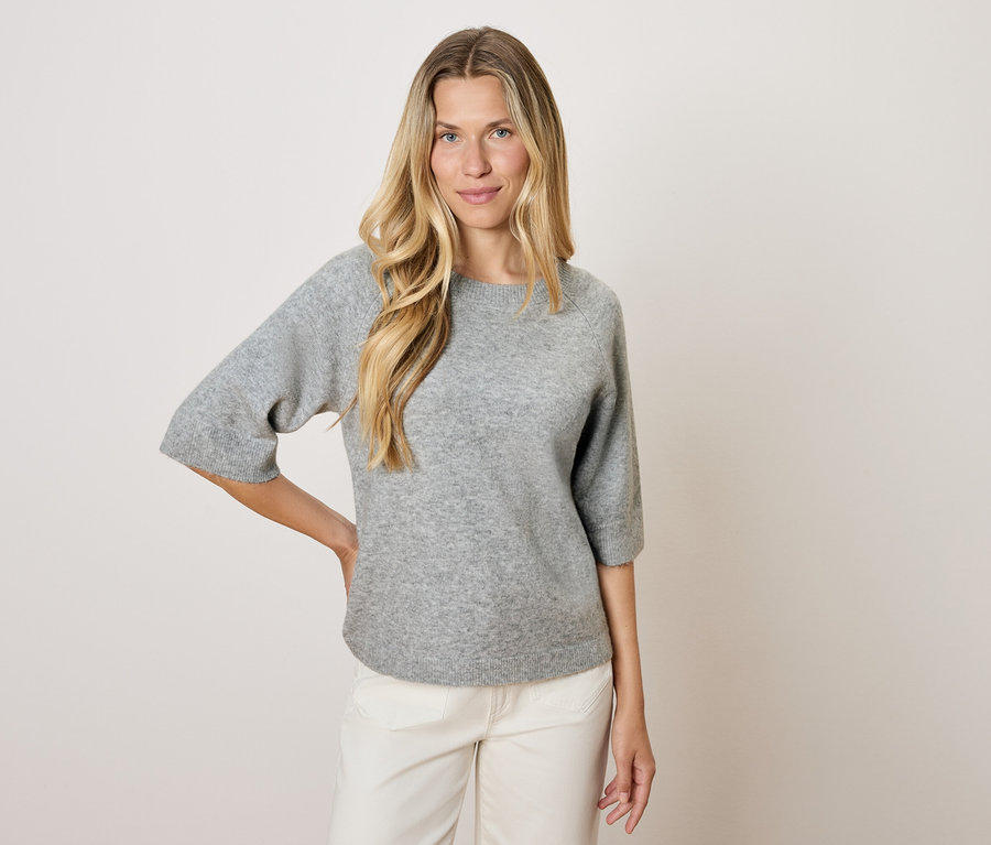 Blondynka w szarym swetrze z rękawami trzy czwarte i białych spodniach pozuje na jasnym tle.