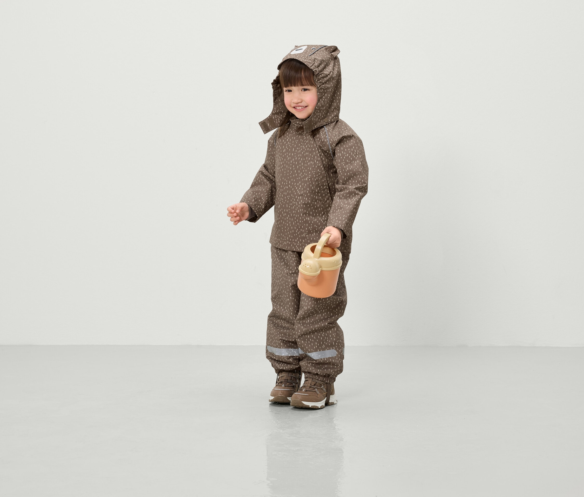 Uśmiechnięte dziecko w brązowej kurtce z kapturem i spodniach w nadruki trzyma pomarańczową konewkę.
