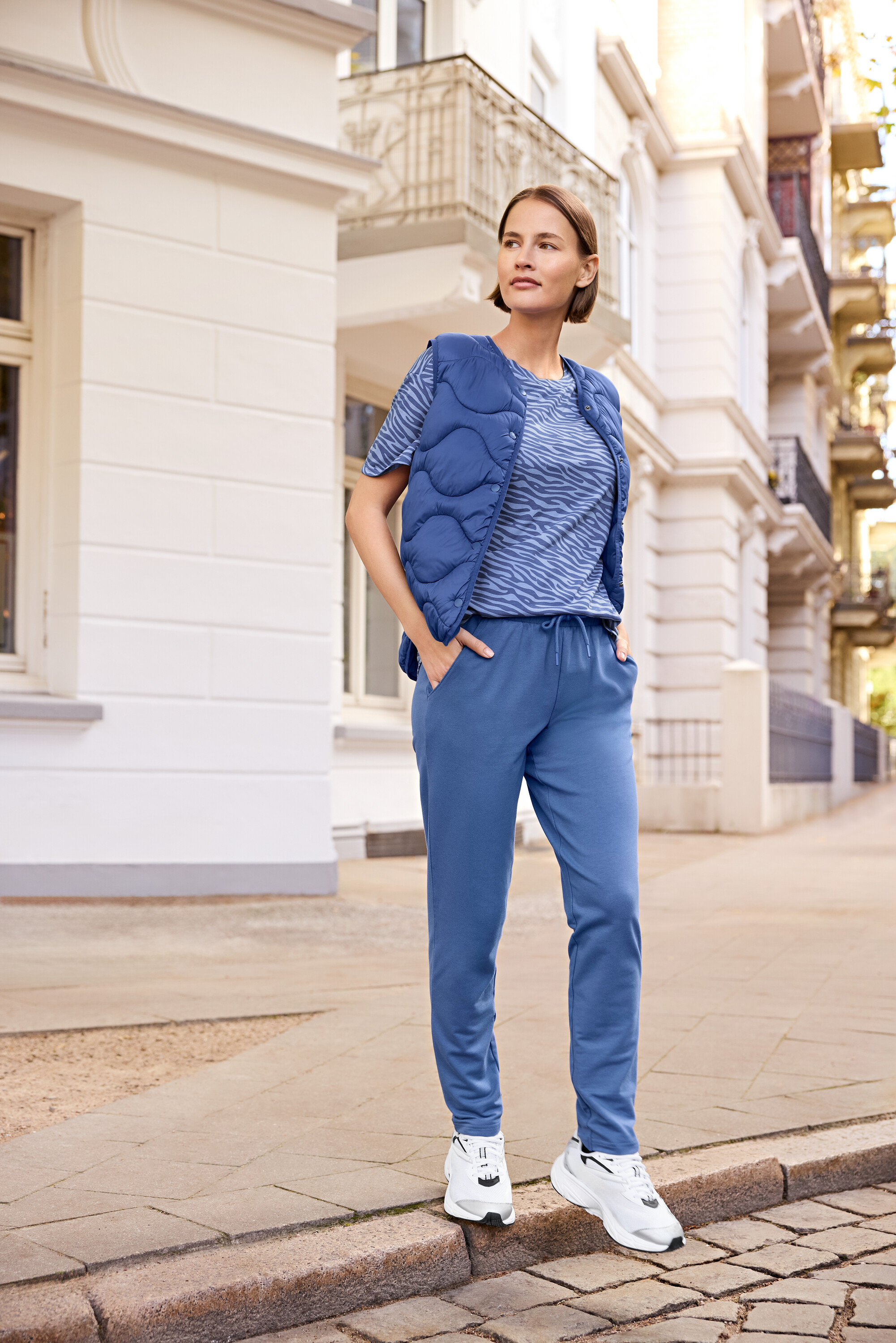 Kobieta stoi na chodniku, ubrana w niebieską kamizelkę, niebieską koszulkę w zebrę i niebieskie spodnie dresowe.