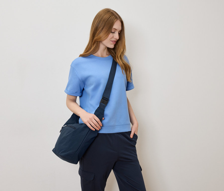 Kobieta z jasnobrązowymi włosami, w jasnoniebieskiej koszulce i granatowej torbie na ramię na kremowym tle.