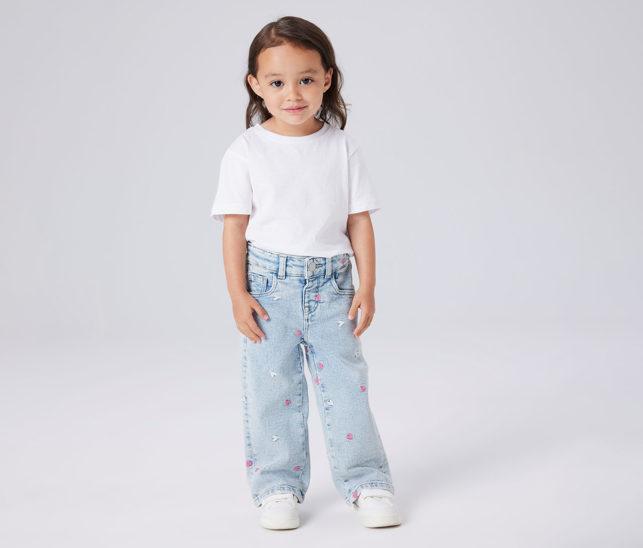 Dziewczynka ma na sobie białą koszulkę i dziecięce jeansy Wide Leg NAME IT z różowymi serduszkami i białymi myszkami.