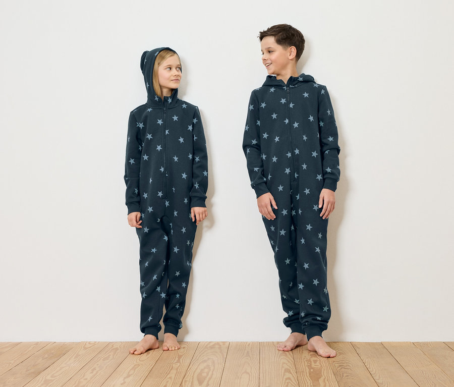 Dziewczyna i chłopak stoją boso na drewnianej podłodze na tle białej ściany, ubrani w granatowe kombinezony w gwiazdki.