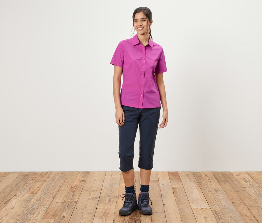 Kobieta stoi na tle białej ściany. Ma na sobie różową koszulę, niebieskie spodnie trzy czwarte i buty trekkingowe.