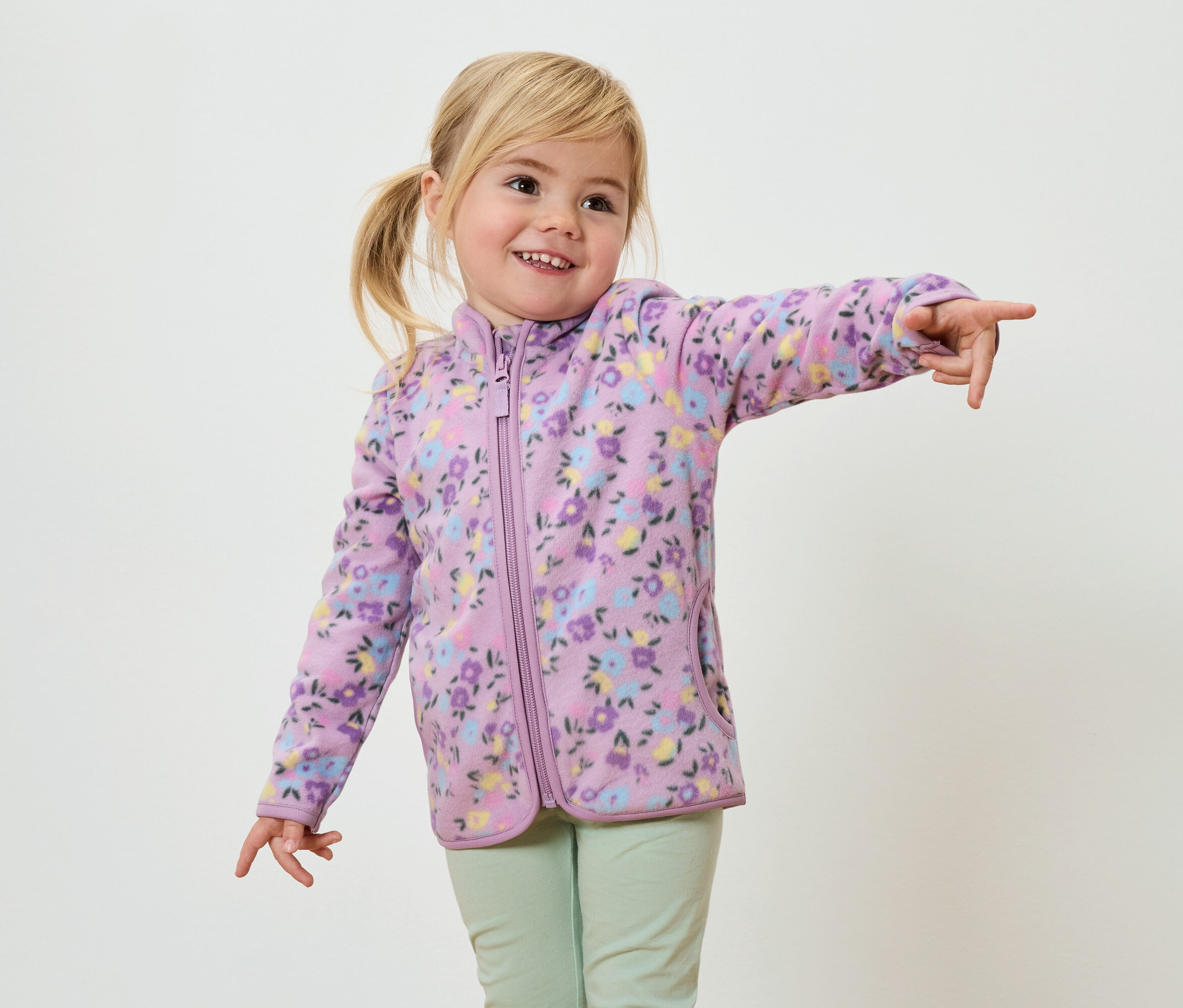 Uśmiechnięta dziewczynka z blond warkoczami, ubrana w fioletową bluzę polarową w kwiaty i jasnozielone spodnie, pokazuje palcem.