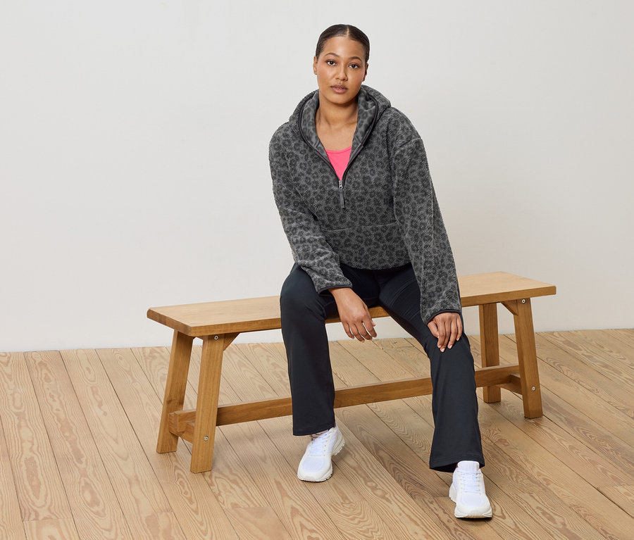 Kobieta siedzi na drewnianej ławce, ubrana w szarą bluzę polarową w lamparcie cętki, różowy top, ciemne spodnie i białe tenisówki.