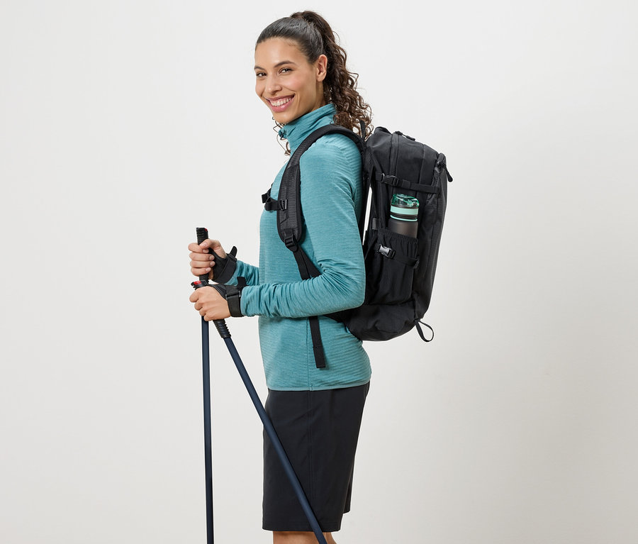 Kobieta w turkusowym golfie, czarnym plecaku i kijkami trekkingowymi.
