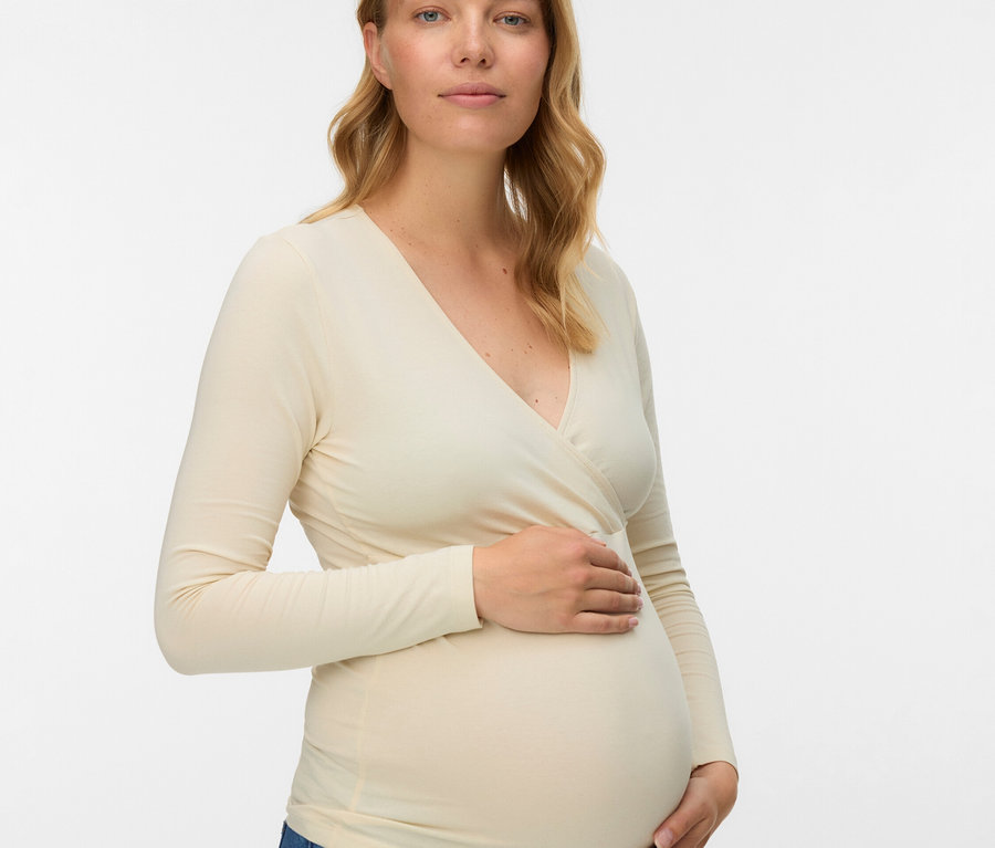 Kobieta w ciąży z blond, falowanymi włosami ma na sobie bluzkę z długim rękawem i dżinsy.