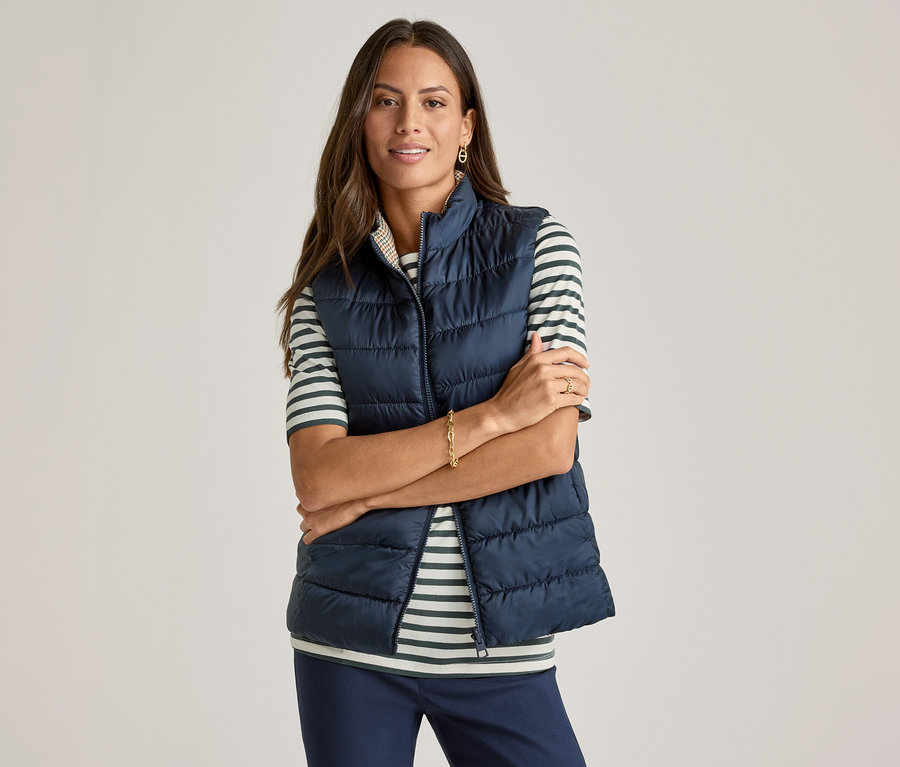 Kobieta ze skrzyżowanymi ramionami ma na sobie granatową kamizelkę nałożoną na koszulkę z półdługim rękawem, w zielone paski.