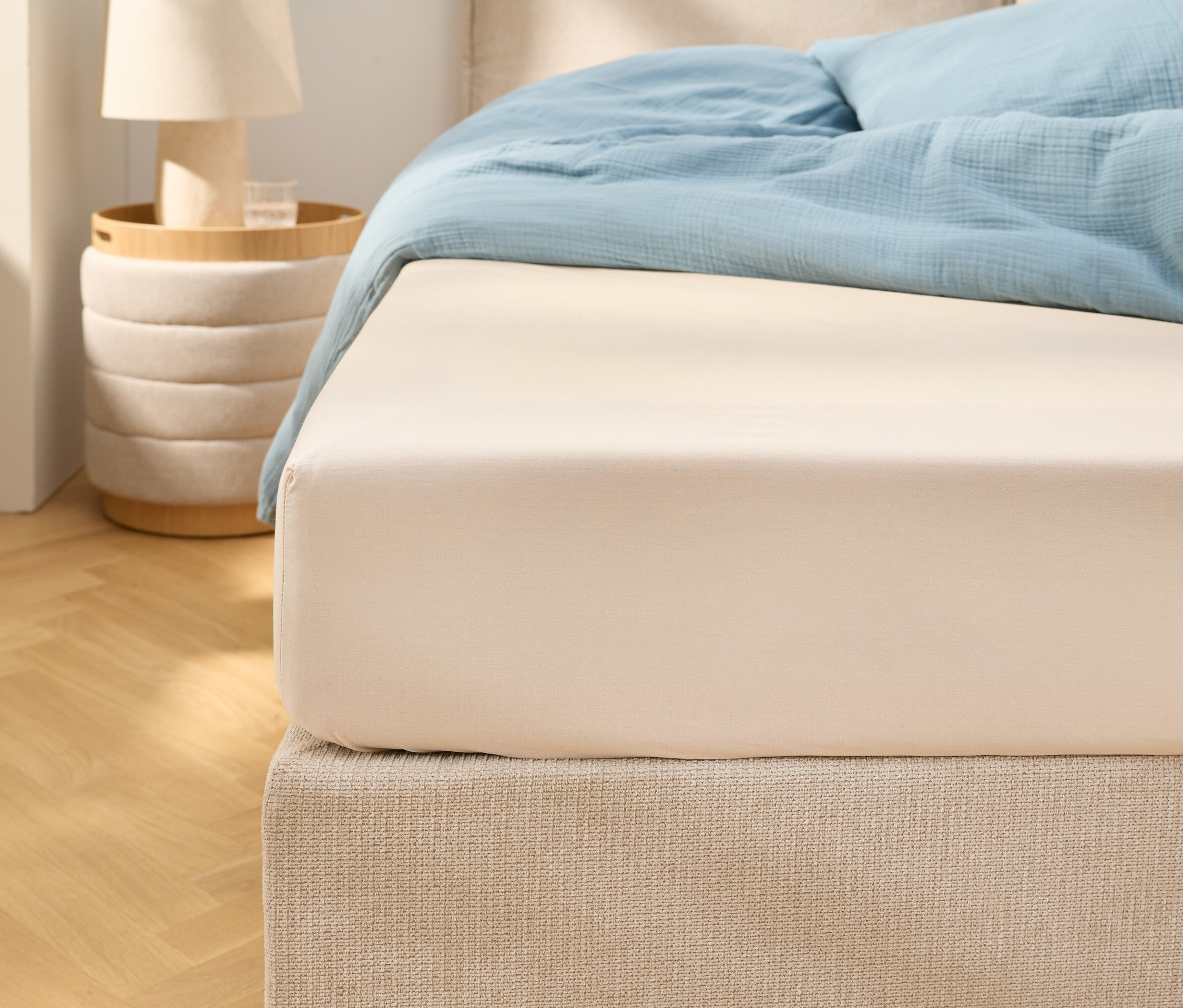 Bliskie ujęcie beżowego prześcieradła z gumką na łóżku z jasnoniebieską pościelą. Obok stoi stolik nocny z lampką.