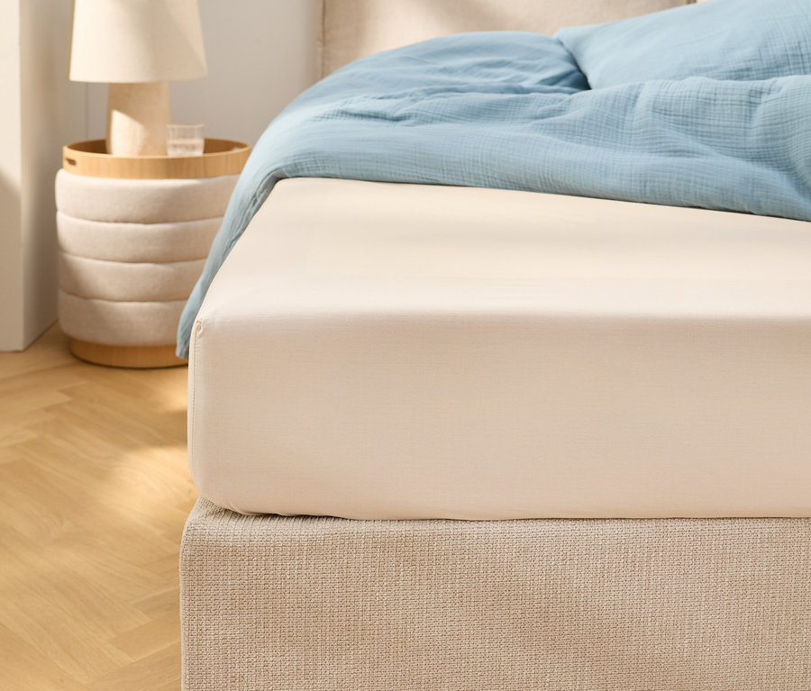 Bliskie ujęcie beżowego prześcieradła z gumką na łóżku z jasnoniebieską pościelą. Obok stoi stolik nocny z lampką.