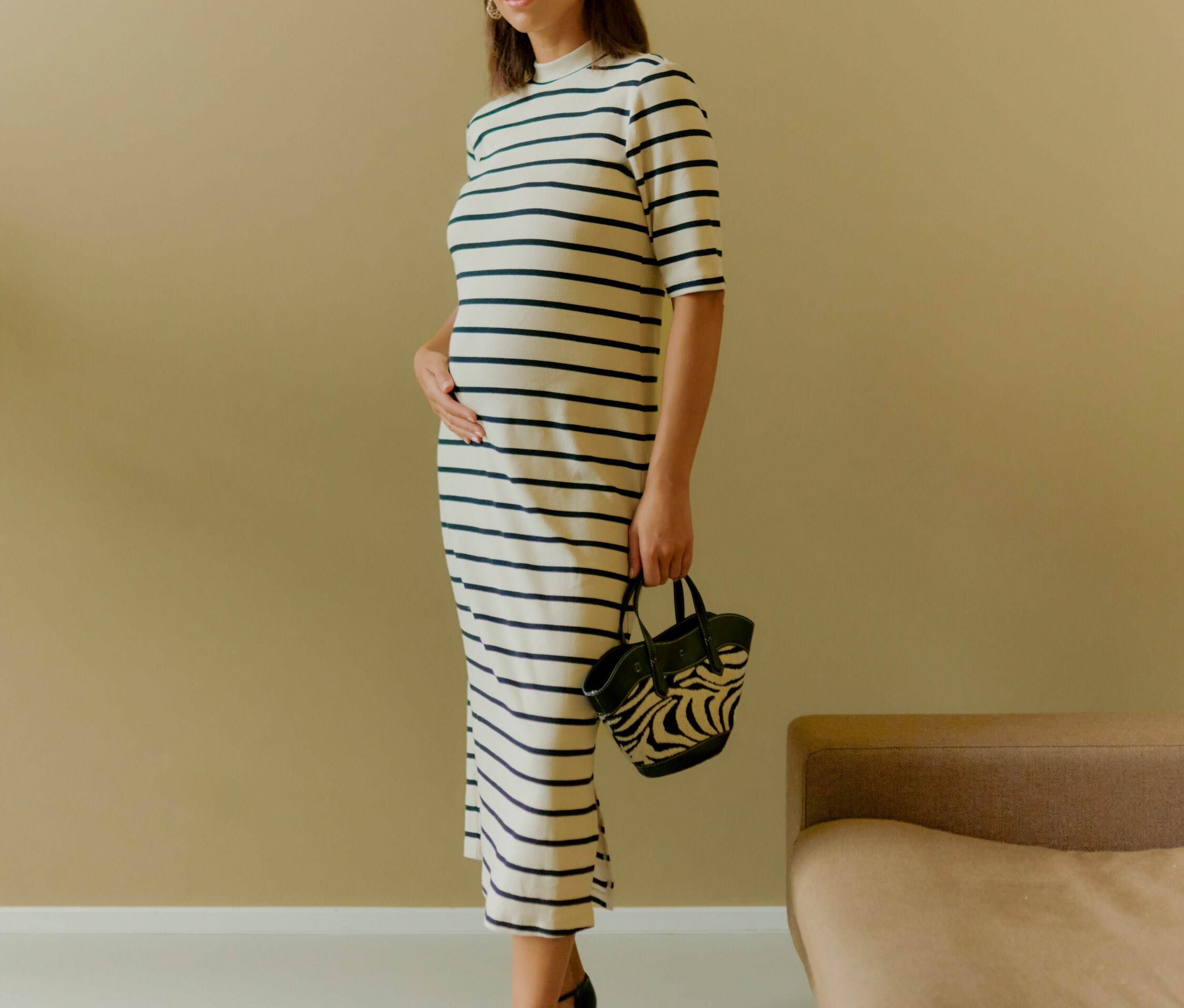 Kobieta stoi i trzyma torebkę w zebrę. Ma na sobie sukienkę w paski i czarne buty.
