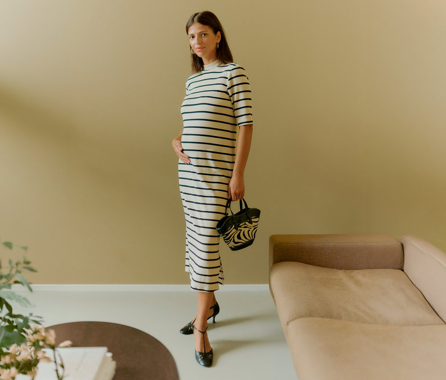 Kobieta w pasiastą sukienkę MAMALICIOUS „MLLANE” trzyma torebkę i dotyka dłonią brzucha. Ma na sobie czarne buty.