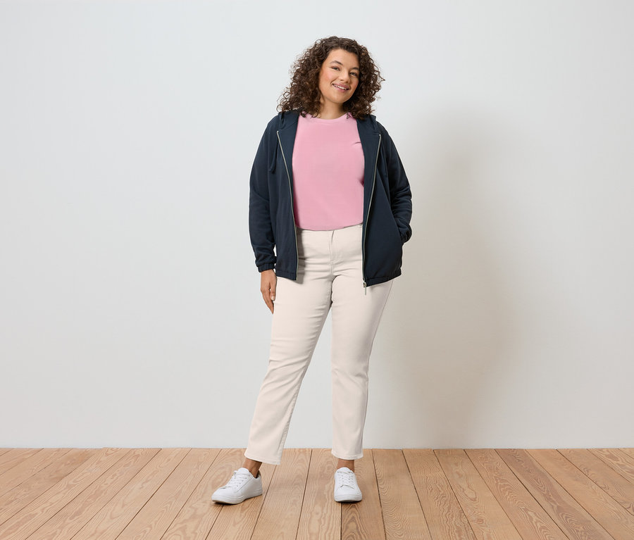 Kobieta o kręconych włosach stoi na tle ściany, ubrana w granatową bluzę z kapturem, różowy T-shirt, kremowe spodnie i białe tenisówki.