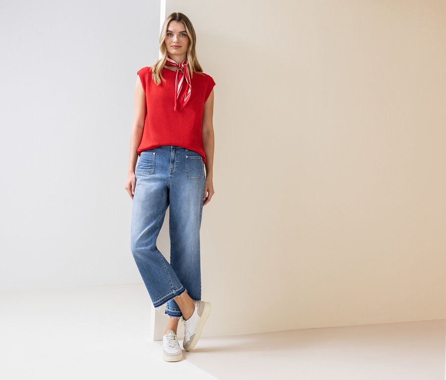 Kobieta stoi i ma na sobie czerwoną bluzkę bez rękawów, apaszkę, dżinsy i tenisówki.