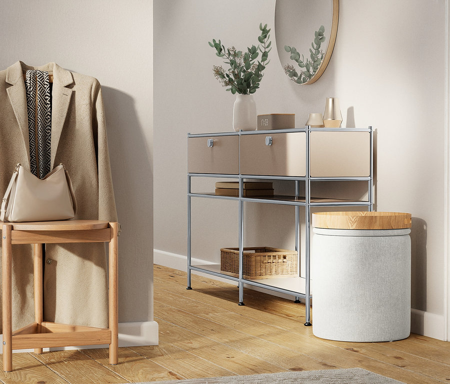 Beżowy płaszcz wisi na ścianie obok stołka z torebką na nim. Obok stoi regał z szufladami i okrągły stołek.