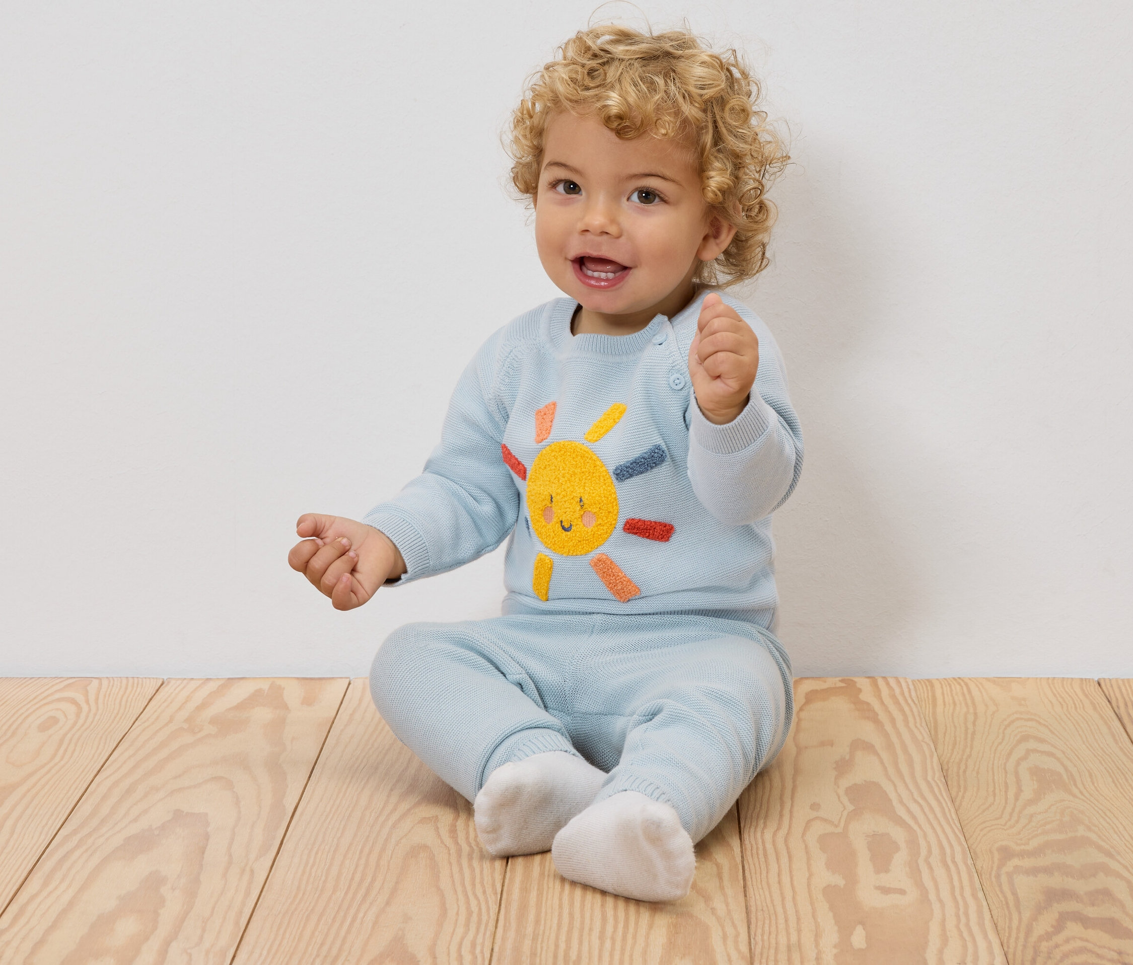 Uśmiechnięte dziecko z blond lokami siedzi na drewnianej podłodze, ubrane w jasnoniebieski sweter z haftowanym słońcem i pasujące spodnie.