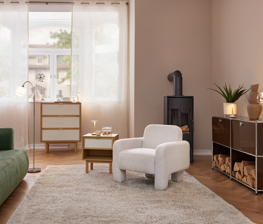 Przytulny salon z zieloną sofą, jasnym fotelem i czarnym piecem na drewno. Komoda i stolik boczny stoją przy oknie z zasłonami. Po prawej brązowy regał z drewnem i lampą stołową.