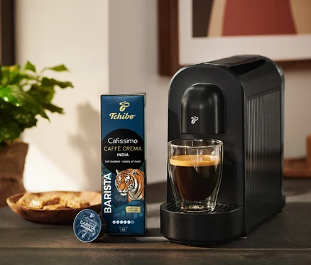 Czarny ekspres do kawy Tchibo Cafissimo ze szklanką kawy, obok opakowanie kapsułek Tchibo Cafissimo Caffè Crema India i jedna kapsułka.