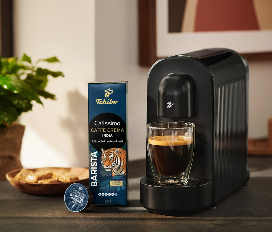 Czarny ekspres do kawy Tchibo Cafissimo ze szklanką kawy, obok opakowanie kapsułek Tchibo Cafissimo Caffè Crema India i jedna kapsułka.