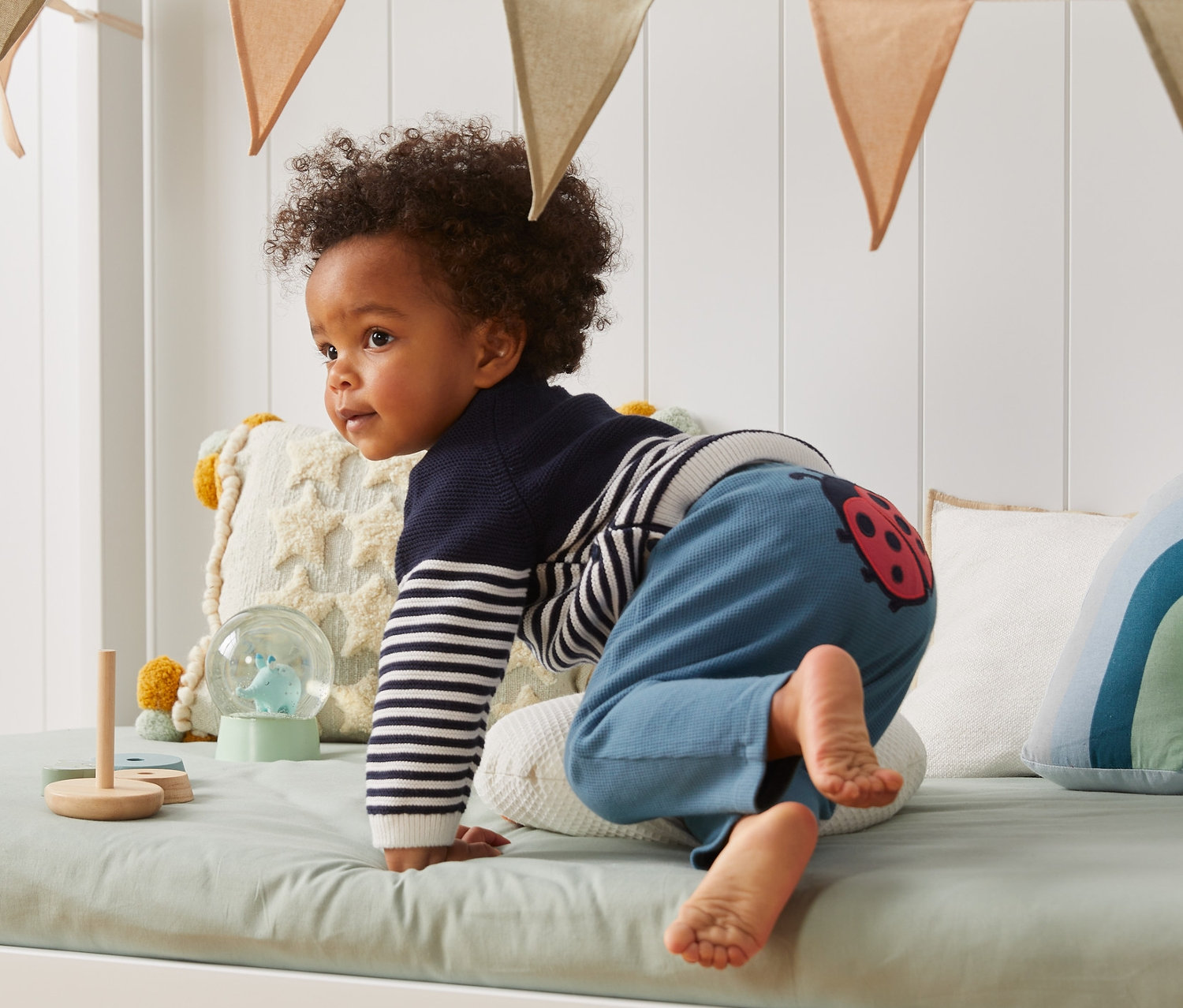 Małe dziecko wspina się na łóżko, ubrane w niemowlęce spodnie dżersejowe.