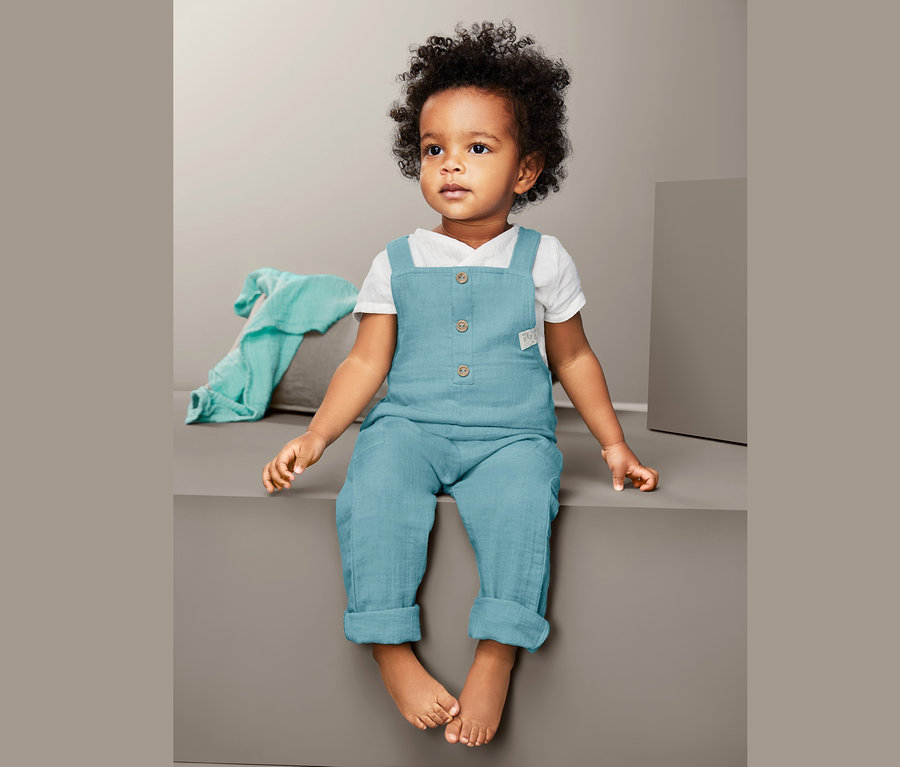 Dziecko siedzi i ma na sobie niebieskie spodnie ogrodniczki.