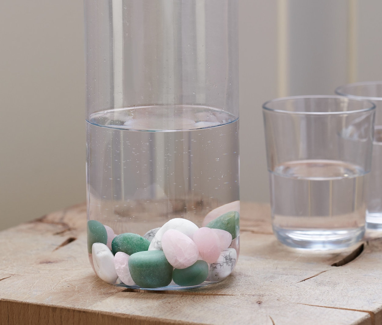 Przezroczysta butelka z korkiem, częściowo wypełniona wodą i Kamienie do wody, stoi na drewnianym bloku obok dwóch szklanek wody.