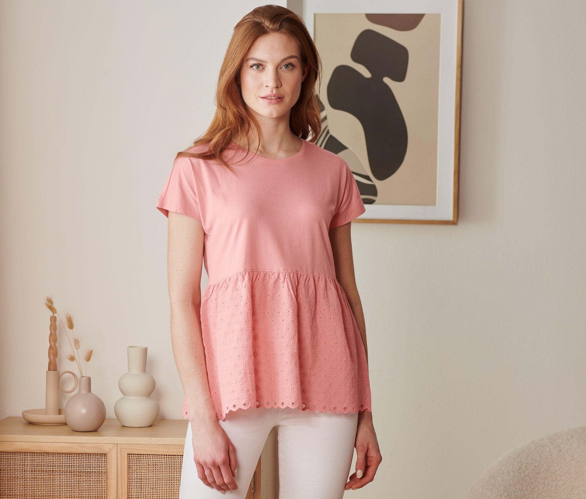 Kobieta ma na sobie koralową bluzkę z mieszanki materiałowej z haftem i białe spodnie.