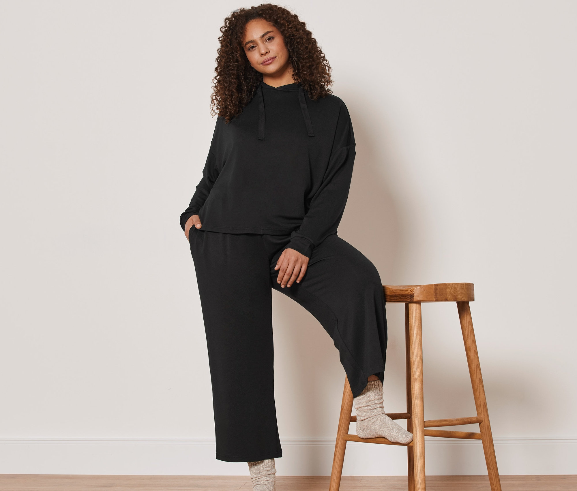 Kobieta pozuje oparta o drewniany stołek, ubrana w czarny dres domowy 7/8.