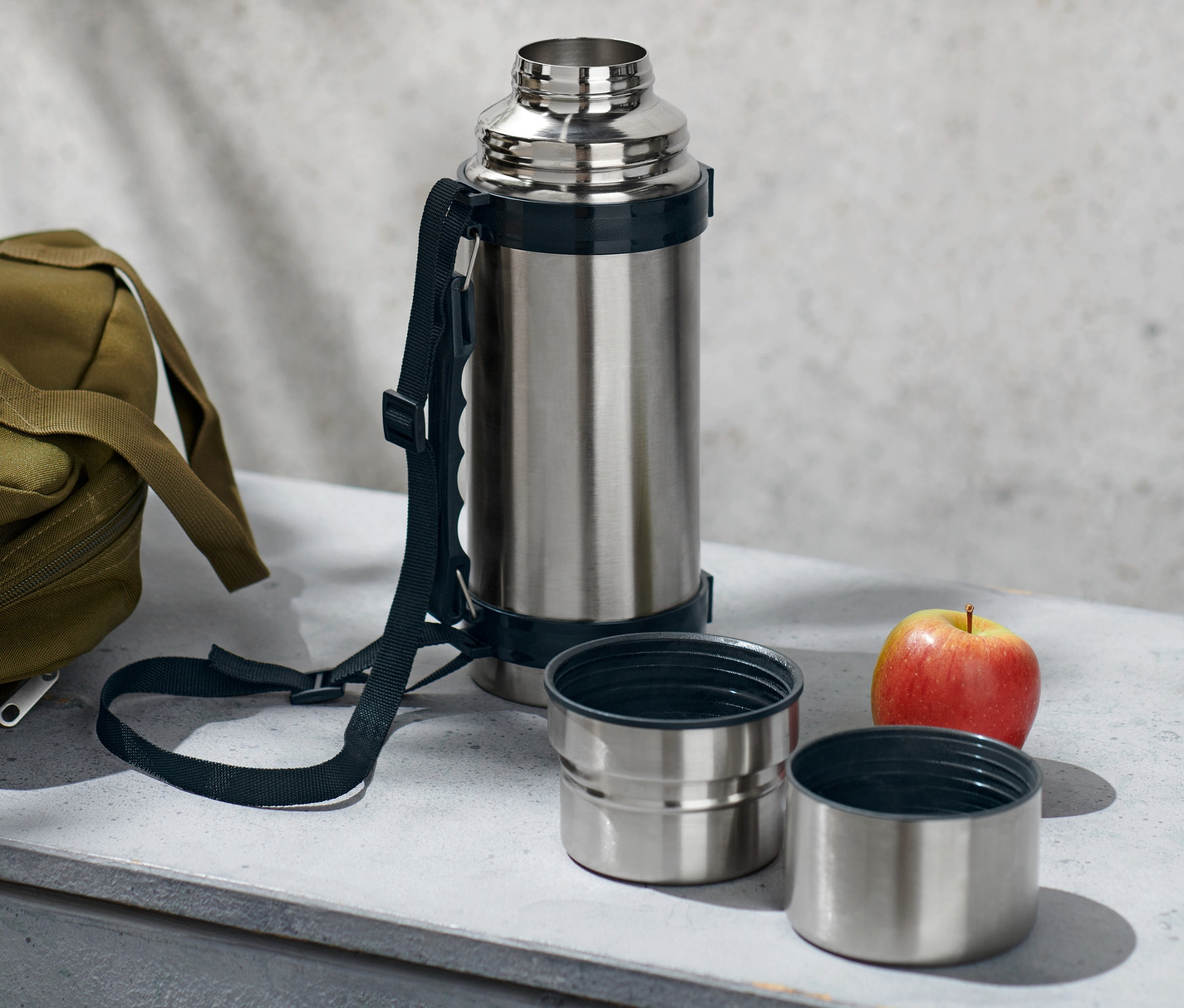 Srebrny termos z dwoma kubkami, oliwkowa torba i jabłko leżą na szarej powierzchni.