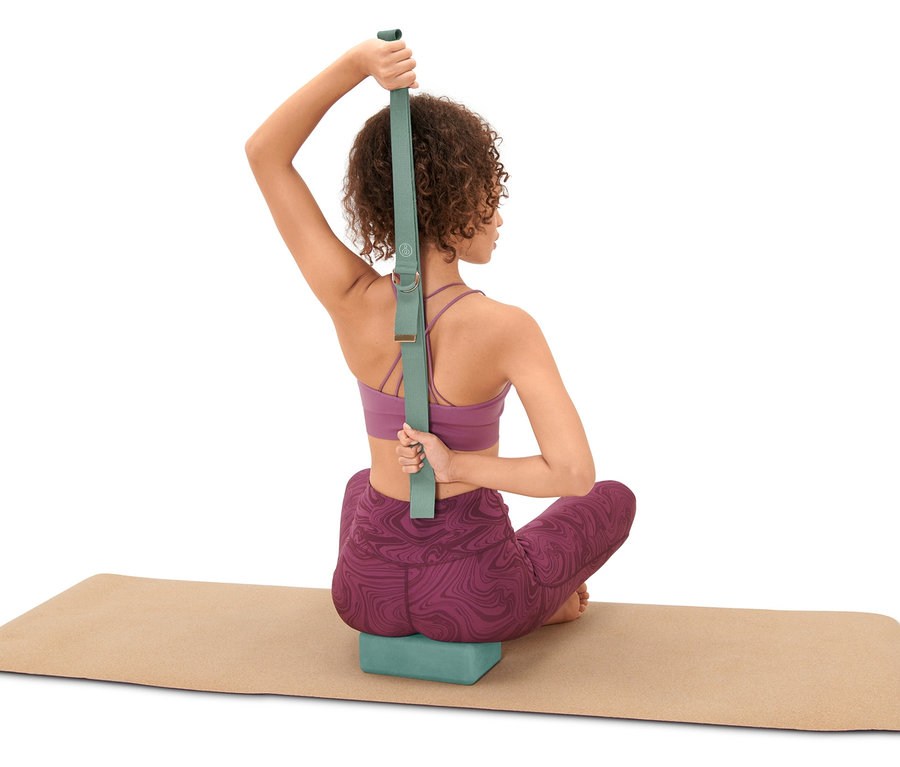 Kobieta siedzi na macie do jogi i używa zestawu do jogi, w tym bloku i paska.