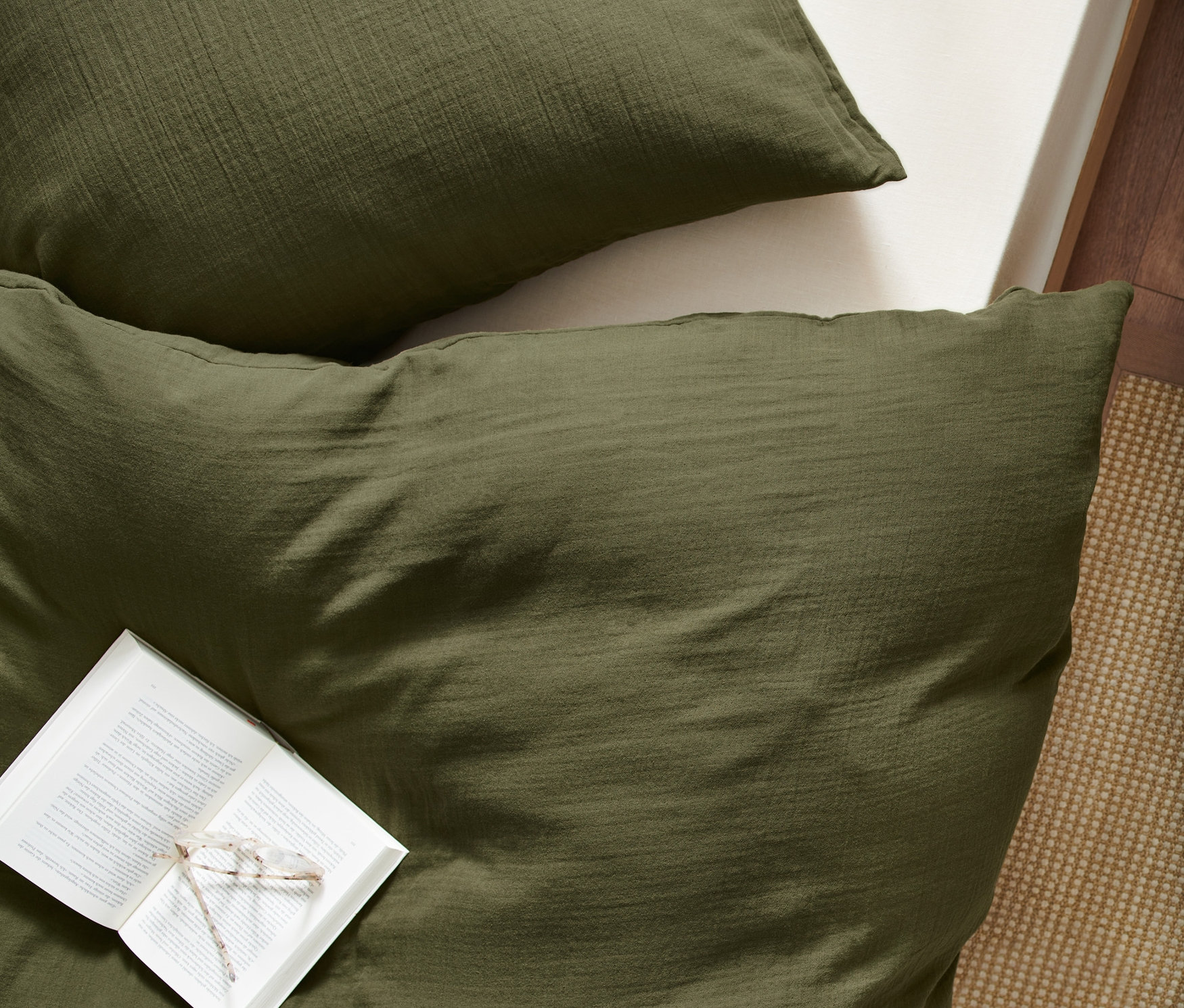 Widok z góry na oliwkowozieloną pościel muślinową w standardowym rozmiarze z otwartą książką i okularami na niej.