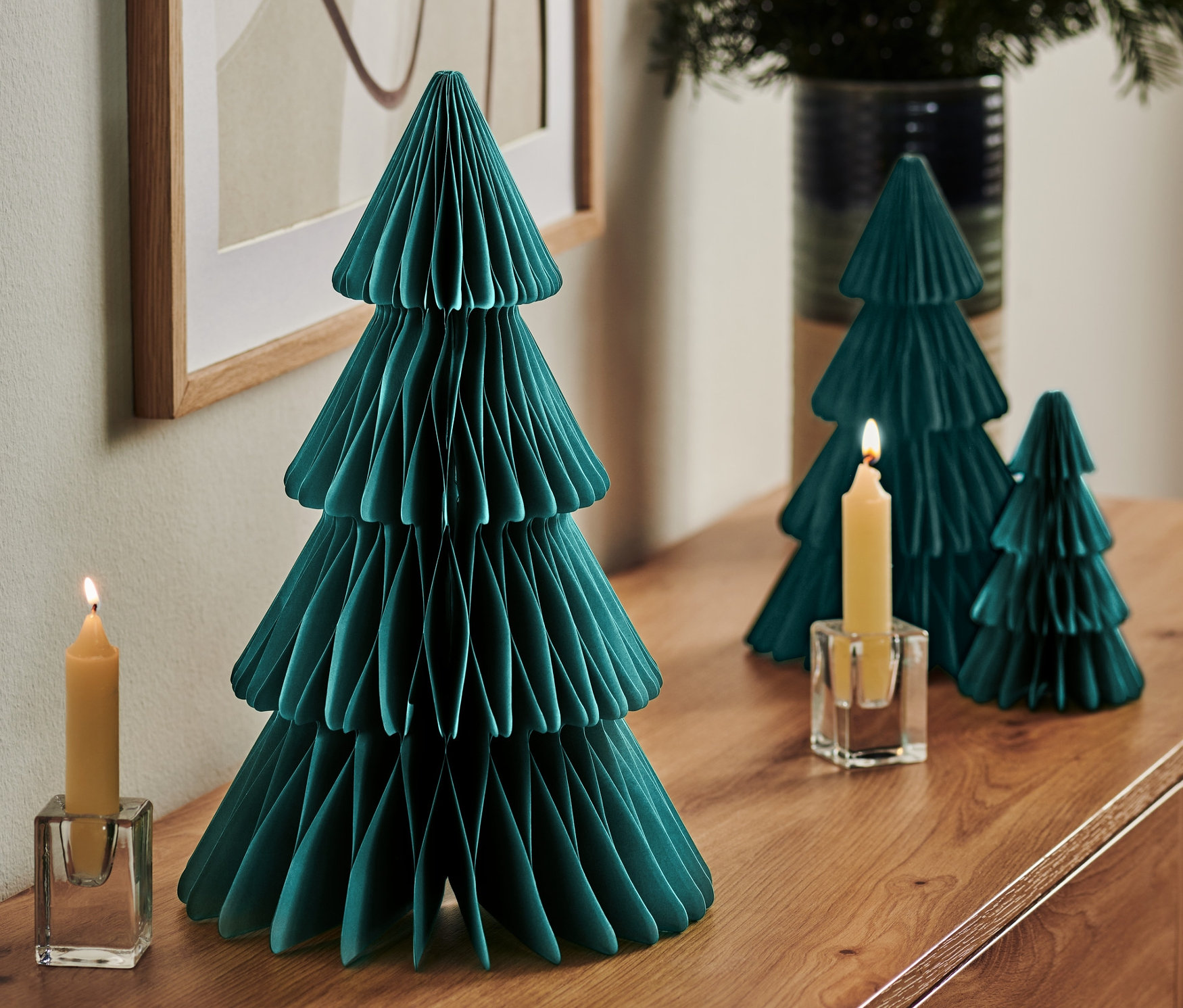 Na komodzie stoi duża składana choinka z papieru, dwie mniejsze choinki, zapalona świeca w szklanym świeczniku i wazon z gałązkami.