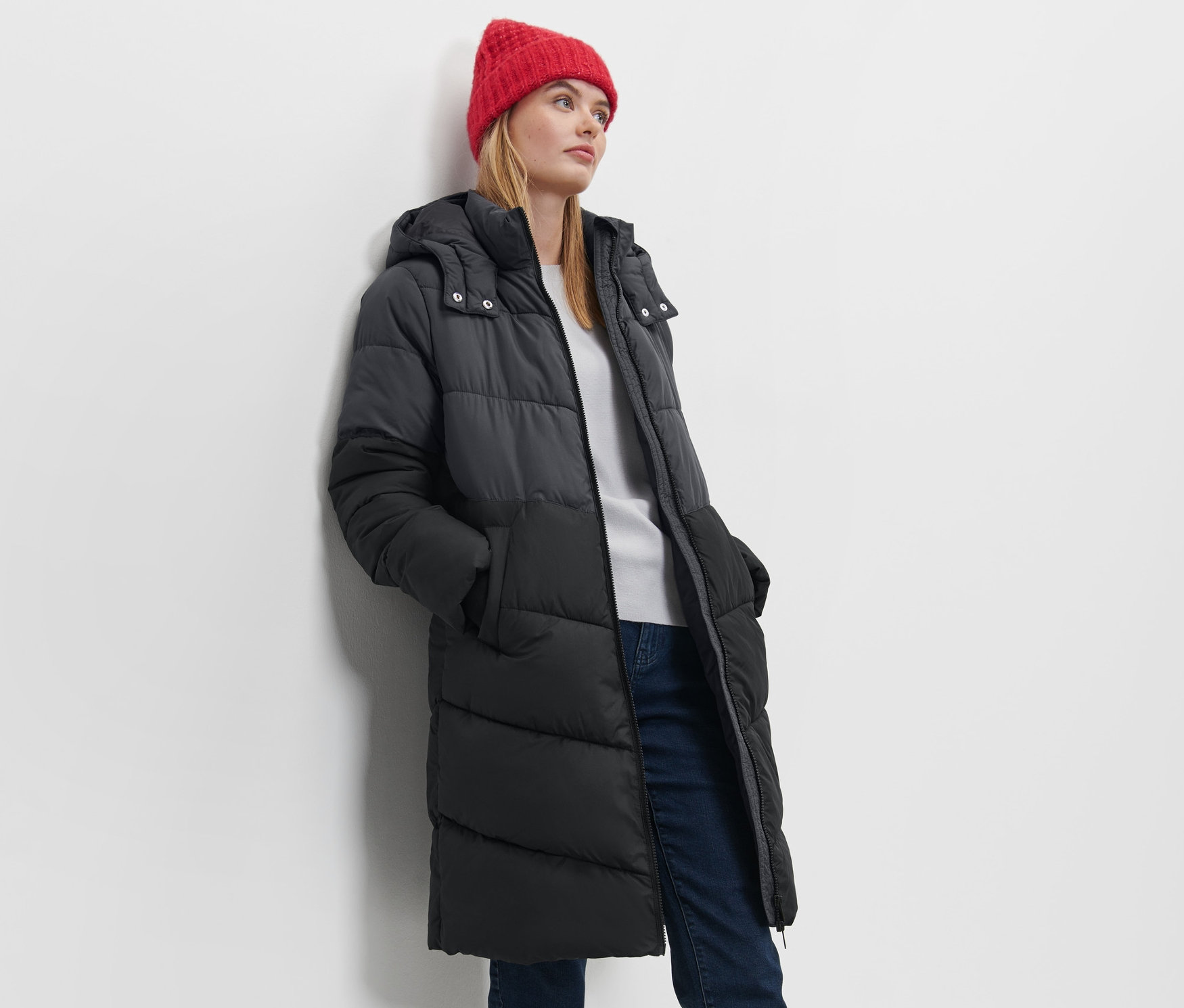 Kobieta pozuje w płaszczu pikowanym z kapturem i czerwonej czapce.