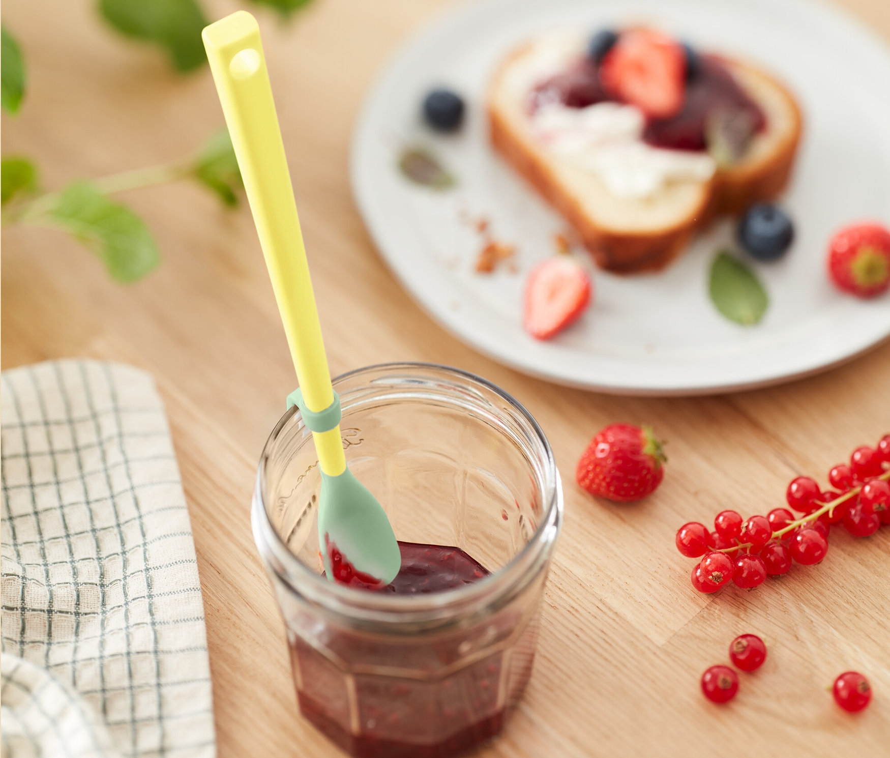 Żółta łyżeczka silikonowa w słoiku dżemu obok talerza z tostem z owocami.