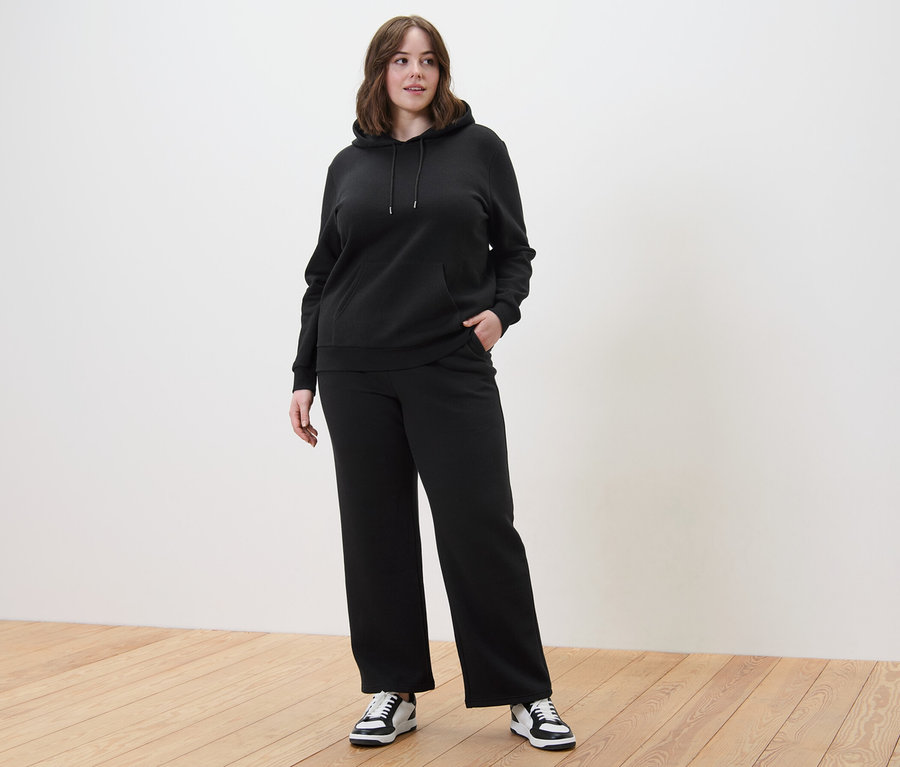 Kobieta w czarnej bluzie z kapturem i spodniach dresowych stoi na drewnianej podłodze.
