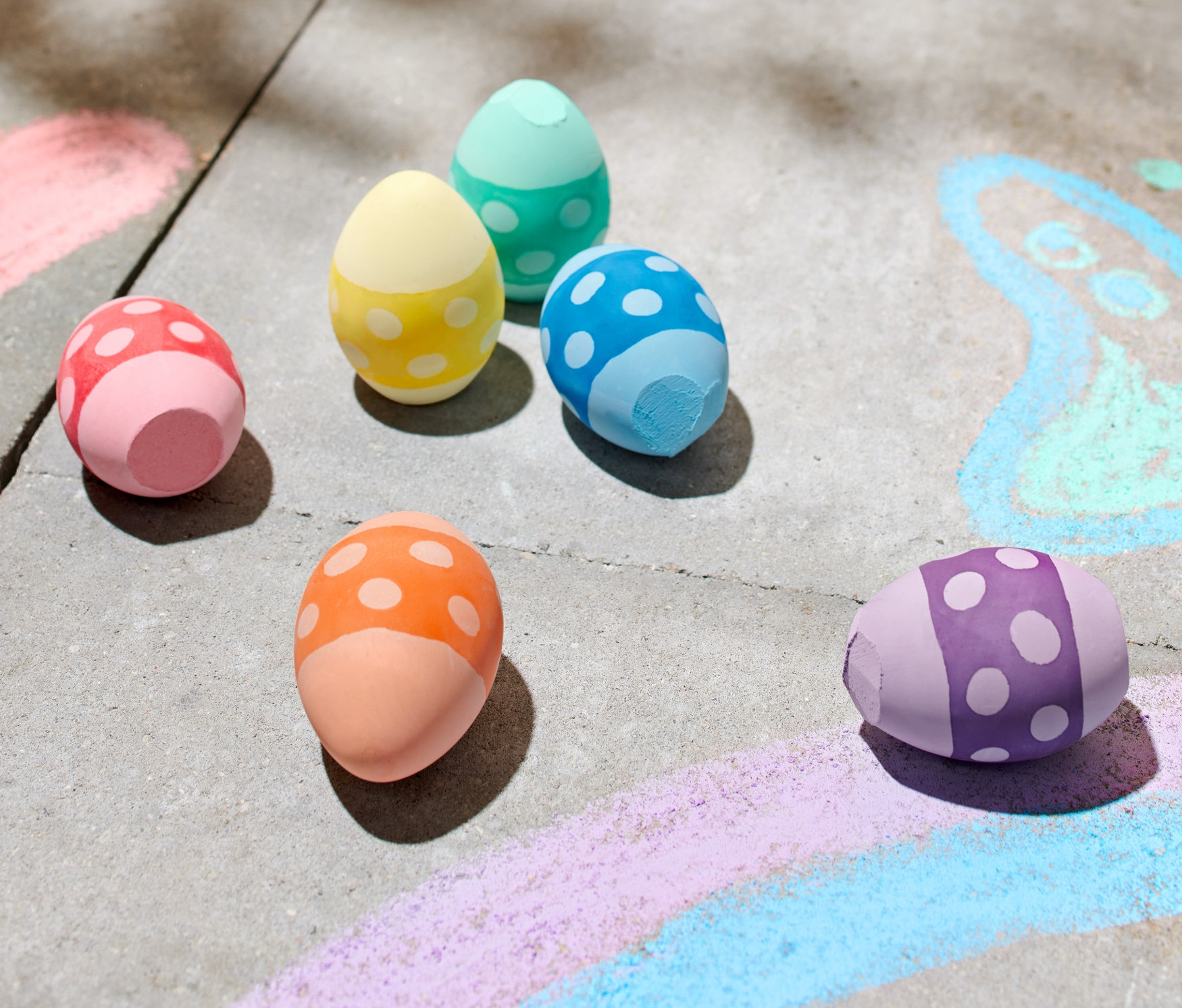 Sześć kolorowych kred w kształcie jajek leży na chodniku obok rysunków kredą.