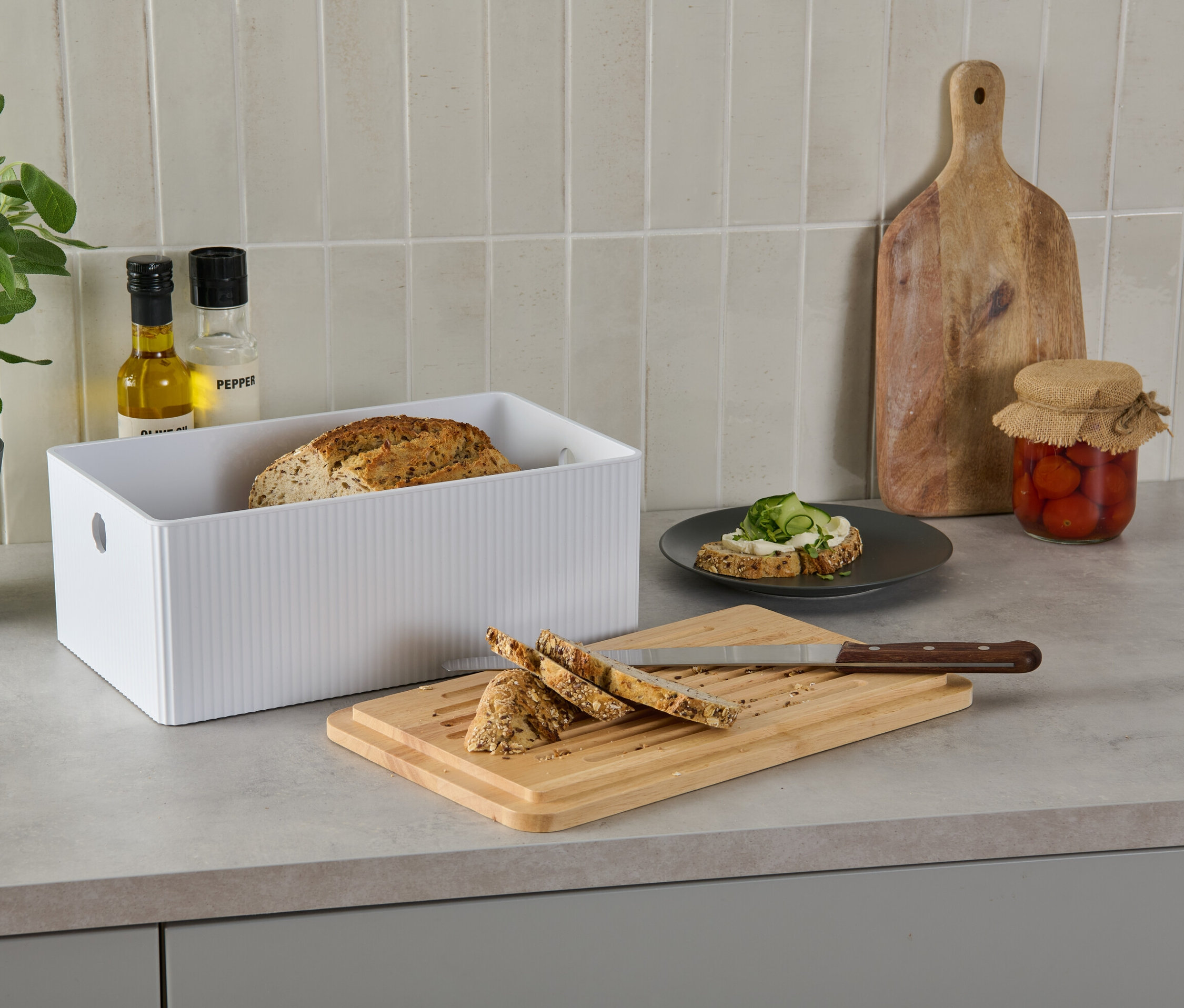 Chlebak z deską do krojenia na blacie kuchennym obok chleba, kanapki, oleju, pieprzu, rośliny i marynowanych pomidorów.