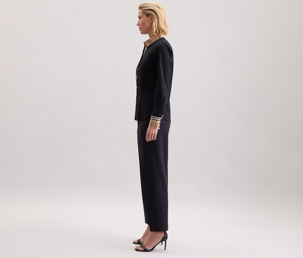 Kobieta z profilu ma na sobie popelinową bluzkę Seidensticker z detalami w kratę, ciemne spodnie i sandały na obcasie.