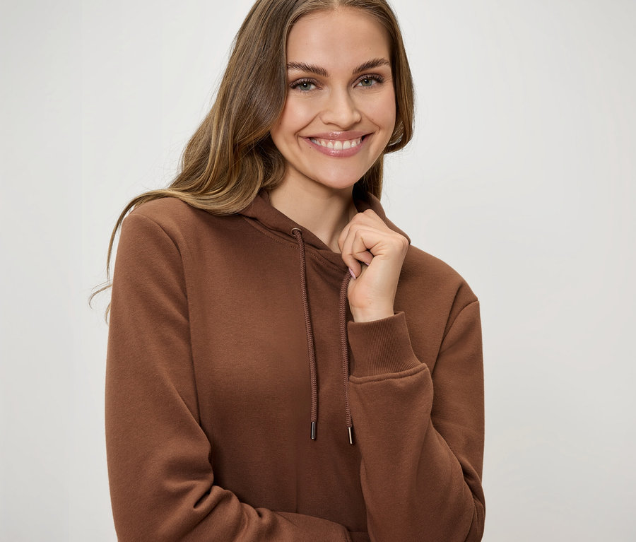 Uśmiechnięta kobieta w brązowej bluzie z kapturem ze sznurkami trzyma jedną ręką kołnierz.