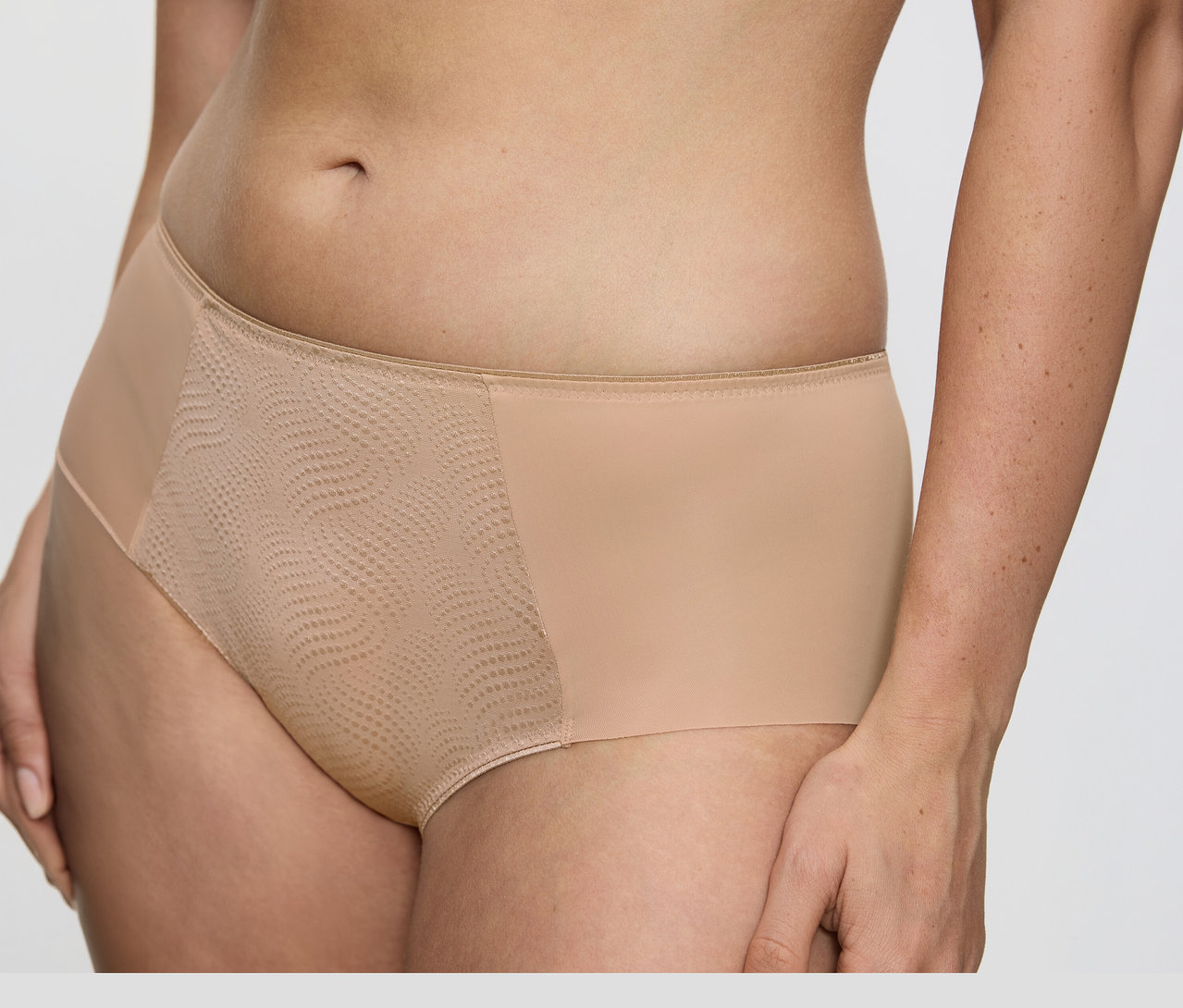 Zbliżenie na tułów osoby ubranej w cieliste figi typu hipster Triumph Essential „Minimizer Hipster X”, z gładkim materiałem po bokach i teksturowanym, wzorzystym panelem z siateczki z przodu.