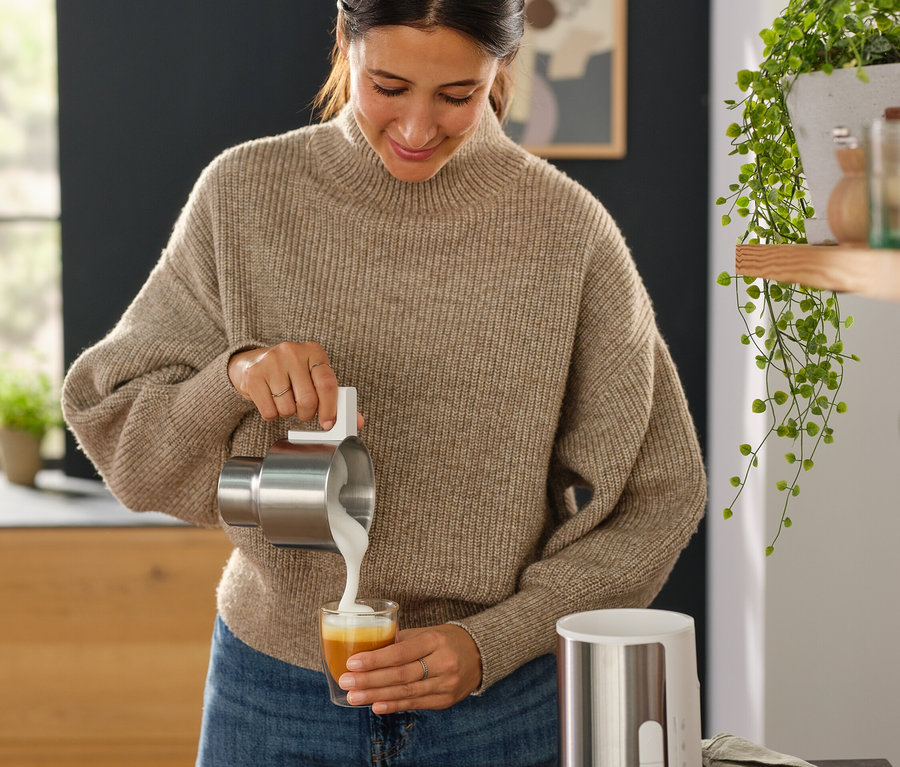 Kobieta nalewa mleko z pojemnika ze stali nierdzewnej do szklanki obok czarnego indukcyjnego spieniacza do mleka.