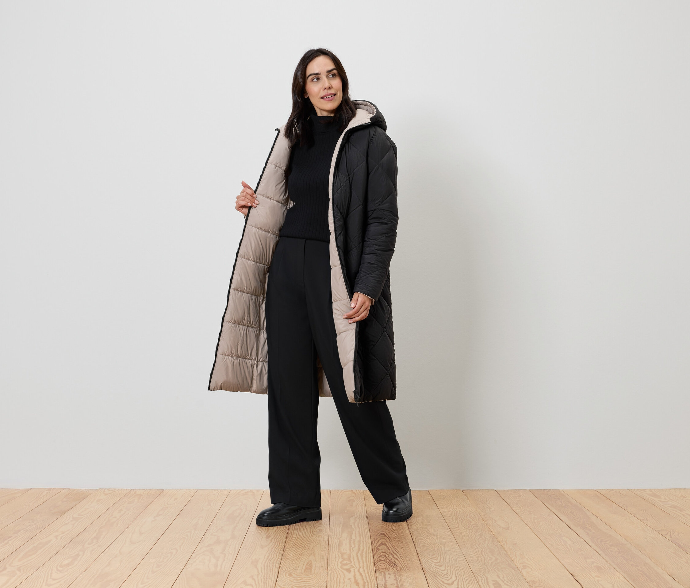 Kobieta pozuje w czarnym płaszczu z beżową podszewką, czarnym golfie i czarnych spodniach.