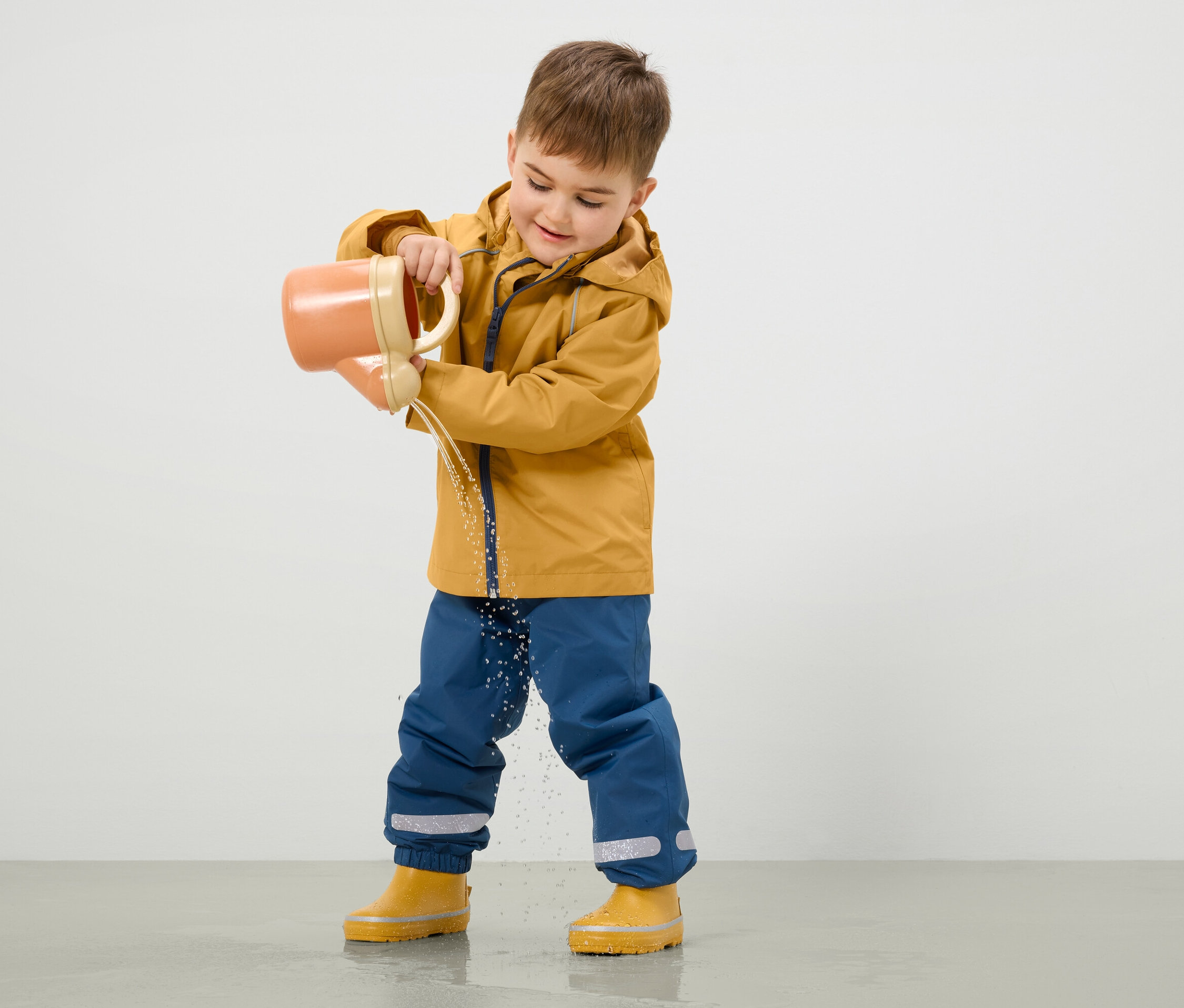 Chłopiec w żółtej kurtce i niebieskich spodniach wylewa wodę z pomarańczowej konewki na ziemię.